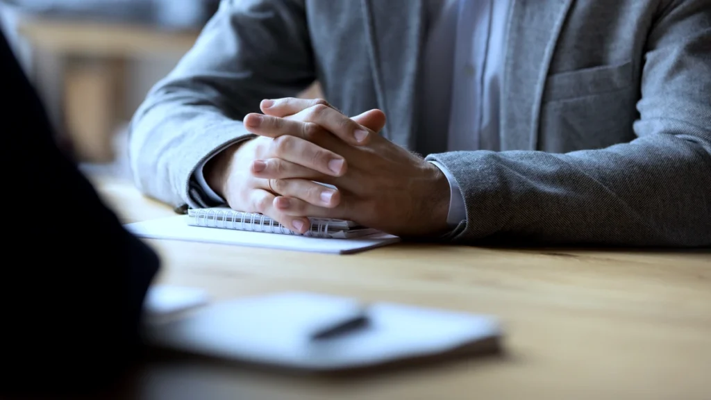Close-up of an attorney with clasped hands at a consultation table, wearing a grey blazer.